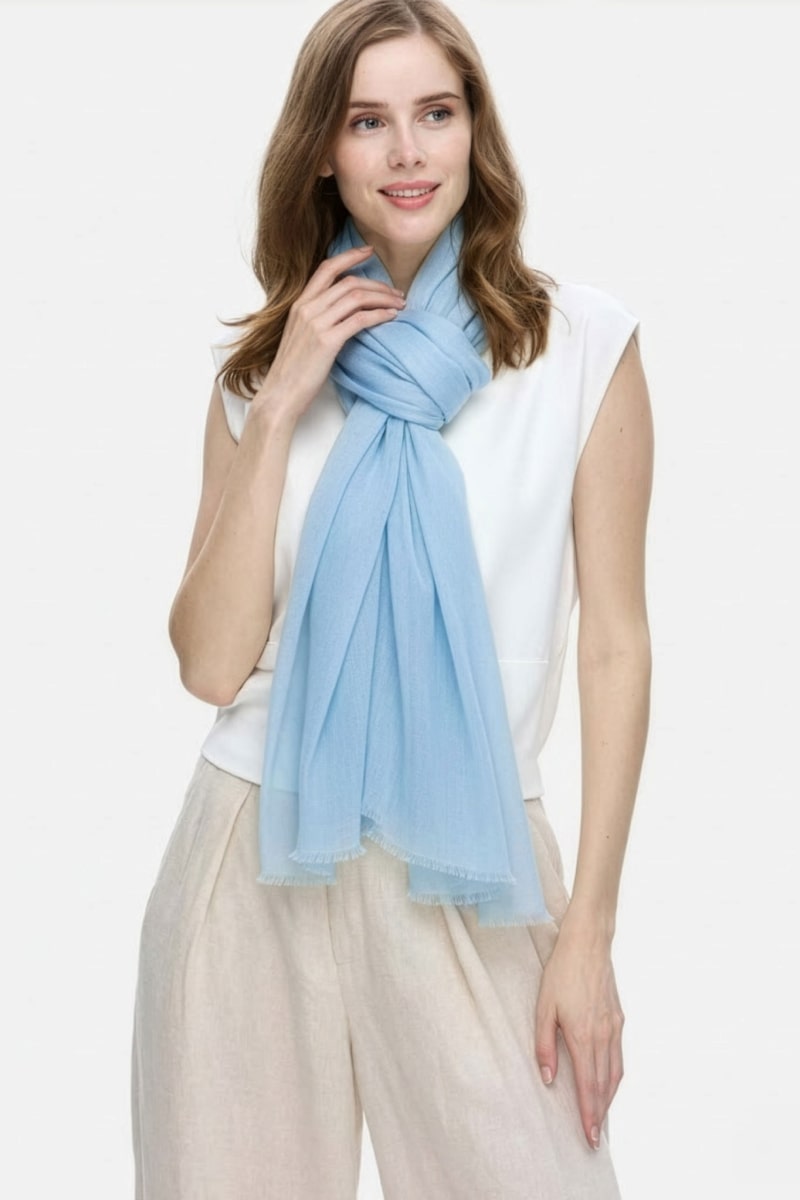 A woman with light brown hair wears a sleeveless white top, beige pants, and the looselyboho Sky Mist 100% Cashmere Scarf draped around her neck. She stands against a white background, smiling softly and gazing to the side.