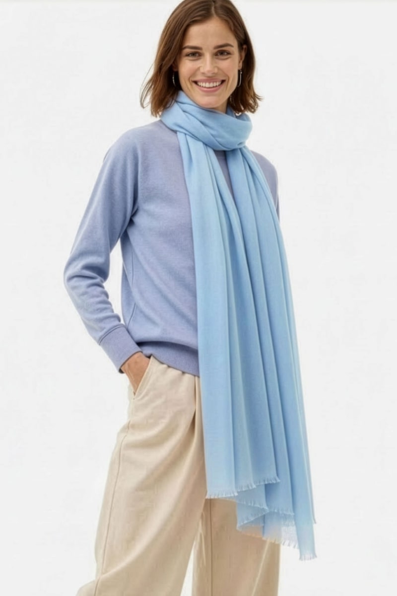 A person wearing the looselyboho Sky Mist 100% Cashmere Scarf in light blue, a light purple sweater, and beige pants stands against a plain white background, smiling with one hand in their pocket.