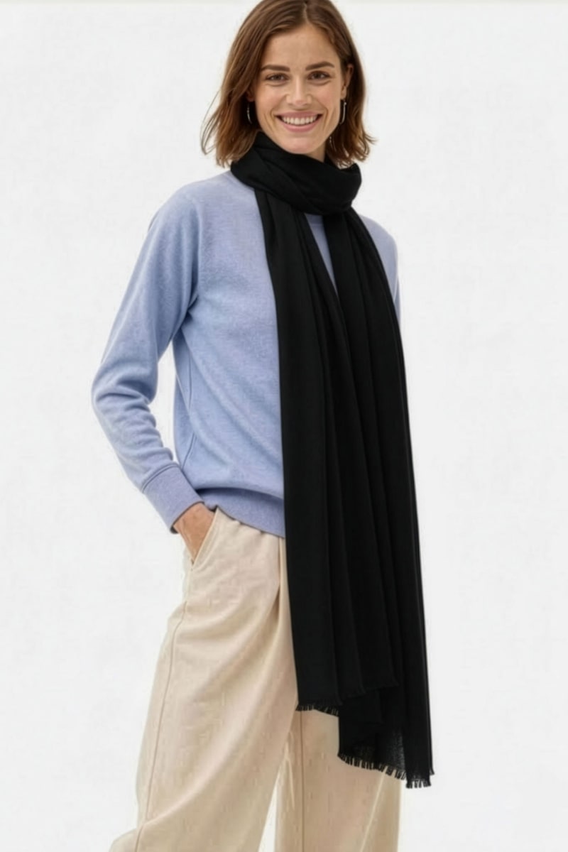 A person smiles with one hand in their pocket, wearing a light blue sweater, beige pants, and the looselyboho Noir Eclipse 100% Cashmere Scarf, posed against a plain white background.