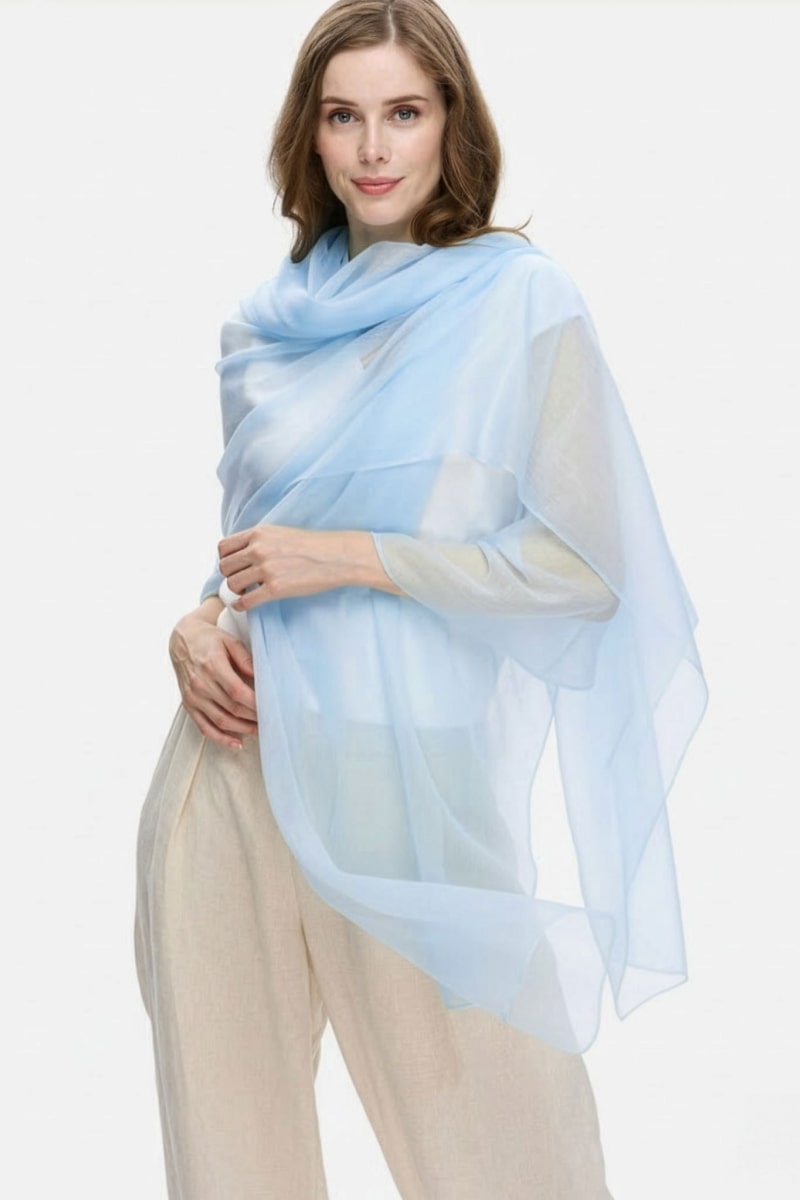 A woman with light brown hair wears the looselyboho Sky Mist 100% Cashmere Scarf, a lightweight light blue accessory draped over her shoulders, paired with a light top and pants, standing against a plain white background.