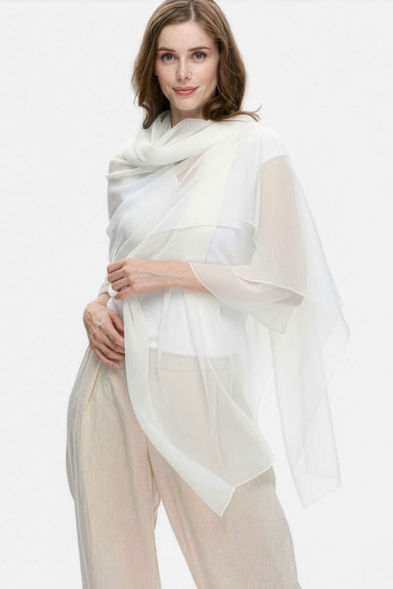 A woman with light brown hair and a slight smile stands against a plain background, elegantly draped in the looselyboho Ivory Whisper 100% Cashmere Scarf over beige pants and a white top, capturing minimalist luxury.