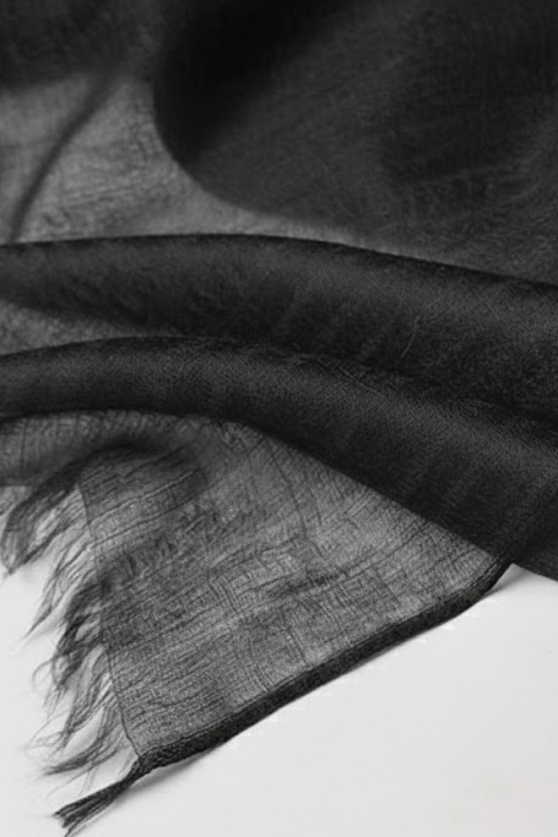 A close-up of the looselyboho Noir Eclipse 100% Cashmere Scarf shows its thin, sheer black fabric with a slightly frayed edge on a white surface, highlighting the delicate elegance of this luxury accessory.