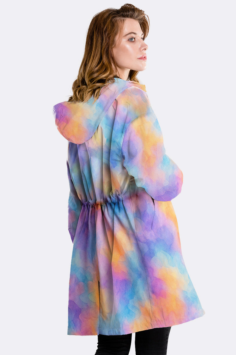 A person with wavy, light brown hair models the looselyboho CANVAS SKY WINDBREAKER—a long, hooded jacket with a pastel tie-dye design and gathered waist—standing turned slightly to the side, facing away.