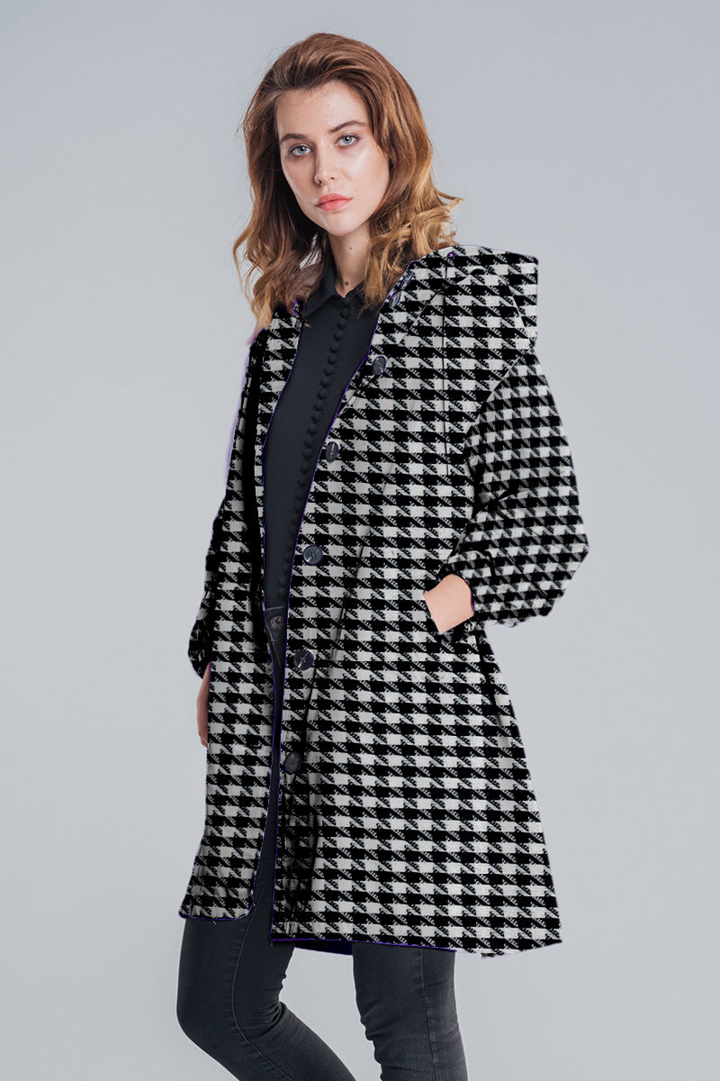 A person stands against a plain background wearing the looselyboho HOUNDSTOOTH WINDBREAKER, featuring a black and white pattern, pockets, and a hood, paired with a black top and dark pants.
