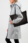 A person wearing a light gray raincoat and black leggings holds "The One" Urban Slate Waterproof Travel Bag by looselyboho, featuring reinforced handles, over their shoulder. The person stands sideways with their hands in the coat pockets against a plain white background.