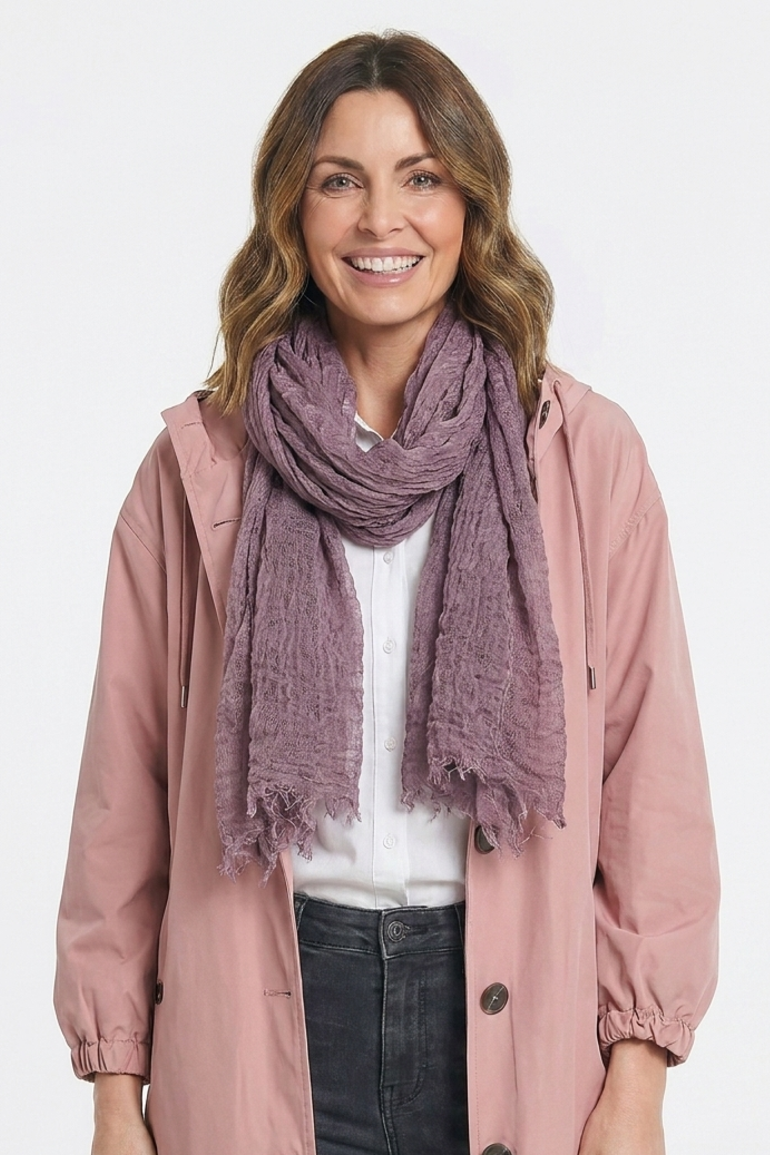 A woman with wavy brown hair smiles in a Lightweight Linen Scarf, styled in mauve, paired with a light pink jacket, white shirt, and black pants. She stands against a plain white background.