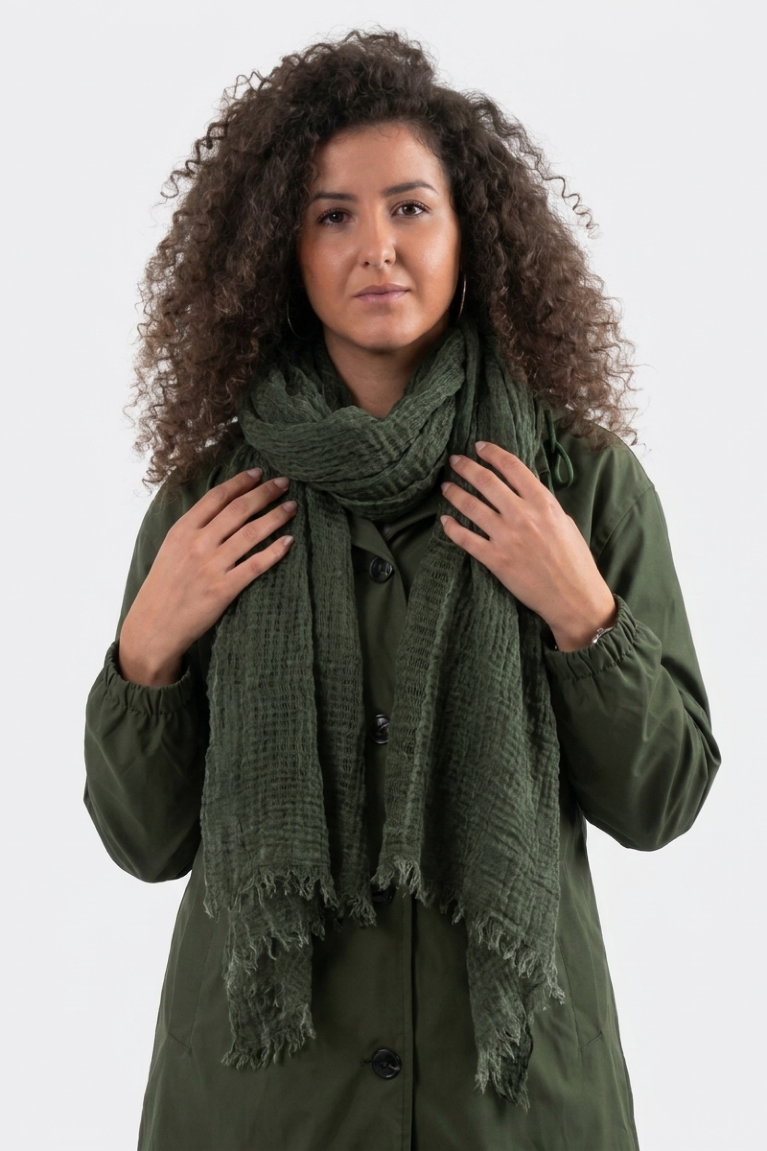 A person with curly hair stands against a plain background, wearing a dark green coat and the Lightweight Linen Scarf, an ultra-breathable accessory, wrapped around their neck. They face forward, hands touching the scarf.