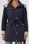 A person is wearing a dark blue lightweight STRIPED NAVY SHADOW WINDBREAKER JACKET from looselyboho with a drawstring waist, white button details, and striped inner cuffs. They are holding a black umbrella in their left hand. The jacket has front pockets and partial snap-button fastening.