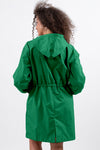 A person wearing The Windbreaker Jacket's EMERALD GREEN WINDBREAKER is seen from behind against a plain white background. The water-resistant material of the jacket features a cinched waist, creating a slight gathered effect. The adjustable hood rests comfortably as the person's curly hair cascades down the back.