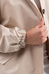 A person wearing a stylish rain-jacket is shown from the shoulders to the waist. They have a bracelet on their wrist and are holding the coat with a hand that has red nail polish. The lightweight, water-resistant BISCUIT BEIGE WINDBREAKER from looselyboho features an elastic cuff and a dark button visible.